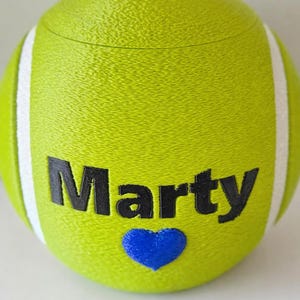 Könnte beinhalten: Ein limettengrüner tennisballförmiger Behälter mit dem Namen "Marty" in schwarzen Buchstaben und einem blauen Herzen. Weiße Streifen verlaufen an den Seiten. Vor dem Behälter liegt eine kleine, silberne Münze.