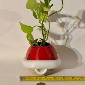 May include: A red and white decorative planter shaped like a Koopa shell, holding a green leafy plant. The planter is approximately 3 inches wide. A yellow measuring tape and a coin are in the foreground.