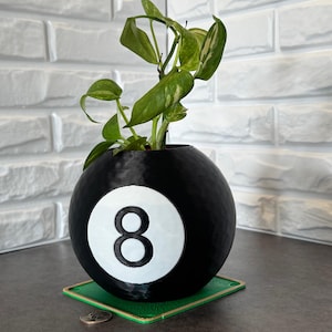 Pode incluir: Um vaso preto e esférico em forma de bola oito, com um círculo branco e o número 8. O vaso contém uma planta verde com folhas verdes claras e amarelas. Está sobre um descanso de copo quadrado verde.