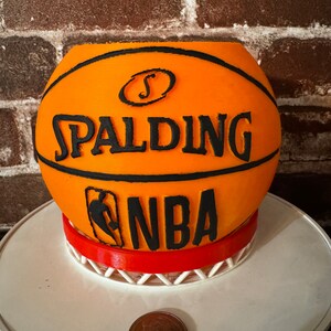 May include: An orange basketball-shaped container with black lettering that reads "SPALDING NBA." The container sits on a red and white base. A quarter is in the foreground. The background is a brick wall.
