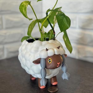 Puede incluir: Una maceta blanca con forma de oveja con textura esponjosa, cara marrón y detalles grises. La maceta contiene una planta verde con hojas. La oveja tiene patas rosas y pequeños cuernos.