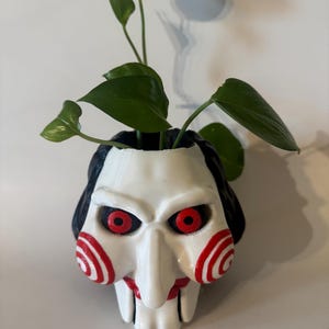 May include: A white and red 3D printed planter shaped like the Jigsaw character from the Saw movie franchise. The planter has red spiral patterns on the cheeks and red eyes. A green plant is growing out of the top of the planter.