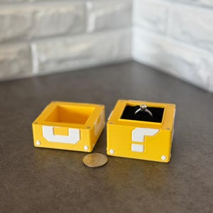 Op de afbeelding: Twee gele en witte ringdozen, één open en één gesloten, met een munt ertussen. De dozen zijn ontworpen om te lijken op het vraagteken en de blok uit de Super Mario Bros. videogame. De gesloten doos bevat een zilveren ring.
