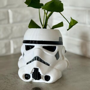 May include: A white Stormtrooper helmet-shaped planter with a black trim and details, holding a green leafy plant. The planter is on a gray surface, with a quarter coin for scale.