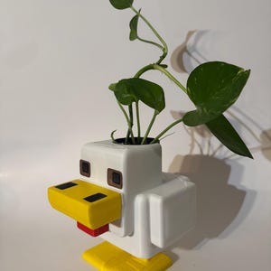 May include: White, yellow, and red 3D printed chicken planter with a green plant growing out of the top. The planter is shaped like a Minecraft chicken.