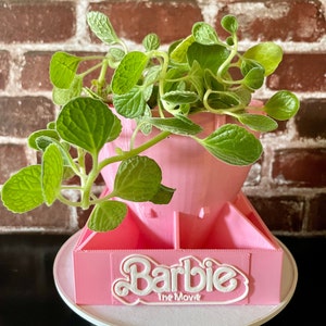 May include: A pink plastic planter with a Barbie The Movie logo. The planter is shaped like a rocket and has a small plant growing out of it.