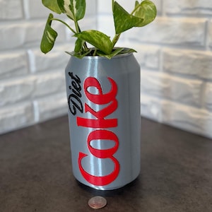 May include: A gray Diet Coke can repurposed as a planter, with the Diet Coke logo in red and black. The can is filled with a green plant with variegated leaves. A penny is at the base of the can.