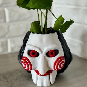 May include: A white and black planter shaped like the Jigsaw Killer's mask, with red eyes and spiral cheeks. The planter holds a green plant with large leaves. A quarter is in front of the planter.