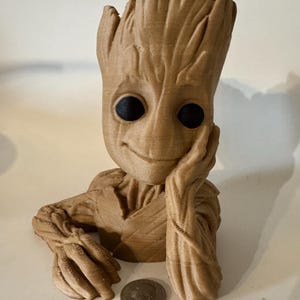 May include: A brown 3D printed figurine of a character from the Guardians of the Galaxy movie, Groot. The figurine is posed with its head resting on its hand, and has a smiling expression. The figurine is about 6 inches tall.