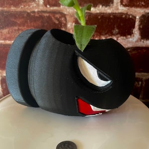 May include: Black and white 3D printed planter shaped like a video game character with a green plant growing out of the top. The planter is sitting on a white surface.