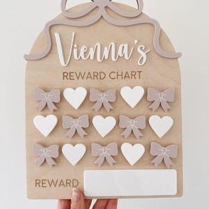 May include: Wooden reward chart with the name "Vienna's" in white script. The chart features a bow design at the top, with spaces for rewards marked by bows and hearts. The word "REWARD" is at the bottom.