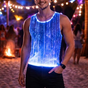 Puede incluir: Una camiseta sin mangas azul de fibra óptica con efecto brillante. La camiseta sin mangas se usa con jeans negros. El fondo incluye una escena de playa con luces de cadena y una hoguera.