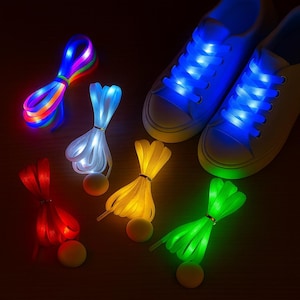 Puede incluir: Zapatillas blancas con cordones azules brillantes, junto a paquetes de cordones LED de colores en rojo, amarillo, verde, blanco y arcoíris. Cada juego de cordones está atado con una pequeña banda dorada y tiene una pequeña batería redonda amarilla.