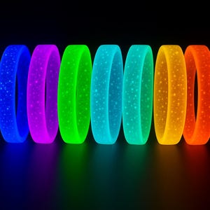 May include: Seven brightly coloured, glowing bracelets arranged in a row. The colours include blue, purple, green, teal, yellow, and orange. Each bracelet has a speckled pattern and is set against a dark background.