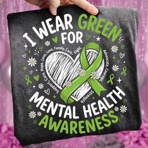 Pode incluir: Camiseta cinza escuro com o texto "I Wear Green for Mental Health Awareness". O design inclui uma fita verde, um coração, margaridas e palavras de apoio como "Amor" e "Esperança".