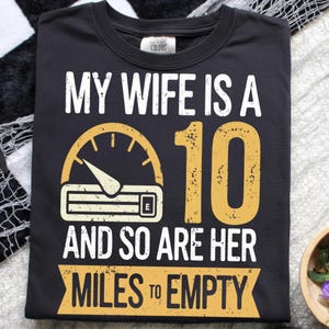 May include: Black t-shirt with white and yellow text. The text reads "MY WIFE IS A 10 AND SO ARE HER MILES TO EMPTY." A speedometer graphic is above the text. The shirt is made of a soft material.