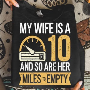 May include: Black t-shirt with white and yellow text: "MY WIFE IS A 10 AND SO ARE HER MILES TO EMPTY." The design features a speedometer graphic. The shirt is held by two hands.