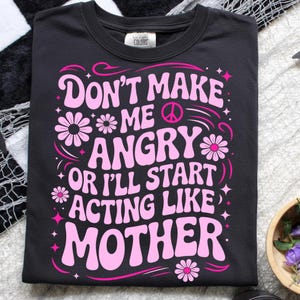 May include: Black t-shirt with a pink graphic that reads "Don't Make Me Angry Or I'll Start Acting Like Mother." The design includes flowers, stars, and a peace sign. The shirt is made of a soft material.