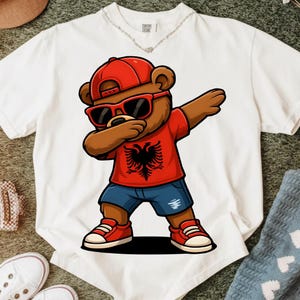 Può includere: T-shirt bianca con un orso dei cartoni animati che fa il dab. L'orso indossa un berretto rosso, occhiali da sole e una maglietta rossa con un'aquila nera. Ha anche pantaloncini blu e scarpe da ginnastica rosse. La maglietta ha il testo "Cotton Colors".