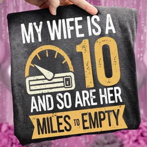 May include: Dark grey t-shirt with the text "MY WIFE IS A 10 AND SO ARE HER MILES TO EMPTY" in white and yellow. The design includes a speedometer graphic.