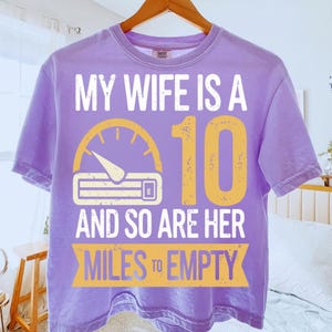 May include: A lilac t-shirt with the text "MY WIFE IS A 10 AND SO ARE HER MILES TO EMPTY" in white and yellow. The design includes a speedometer graphic.