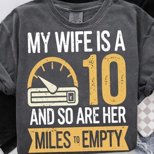 May include: Dark grey t-shirt featuring the text "MY WIFE IS A 10 AND SO ARE HER MILES TO EMPTY." The design includes a yellow speedometer graphic and the number 10.