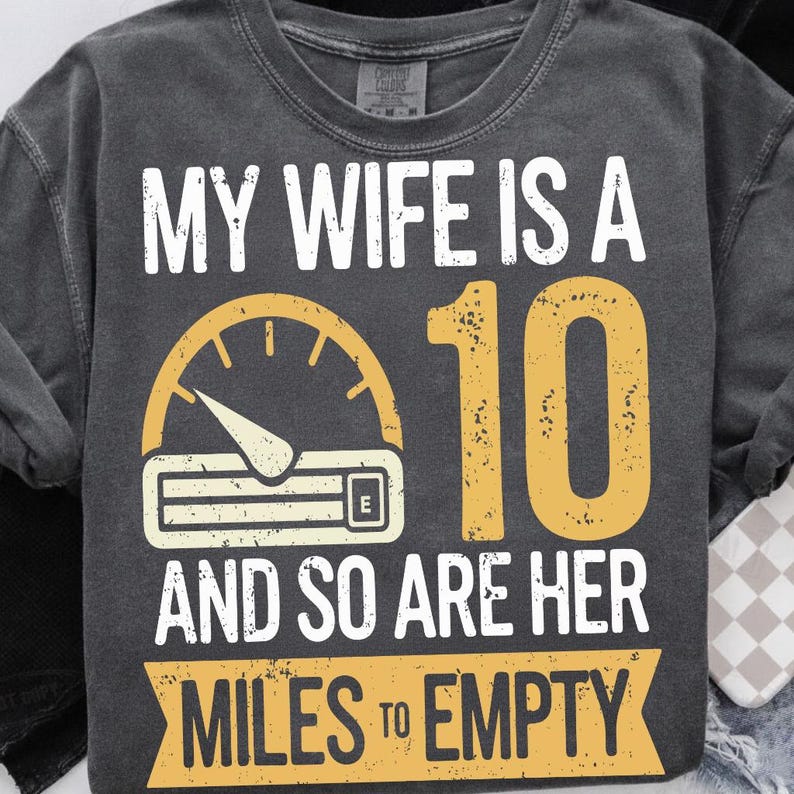 May include: Dark grey t-shirt featuring the text "MY WIFE IS A 10 AND SO ARE HER MILES TO EMPTY." The design includes a yellow speedometer graphic and the number 10.