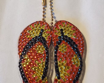 May include: A keychain featuring a pair of red, yellow, and black flip flops with rhinestones. The flip flops are attached to a gold chain and a gold clasp.