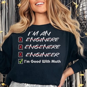 May include: Black long-sleeved sweatshirt with the text "I'M AN ENGINEER" in white. The word "ENGINEER" is checked off with a green tick. The sweatshirt is paired with a silver sequin skirt.
