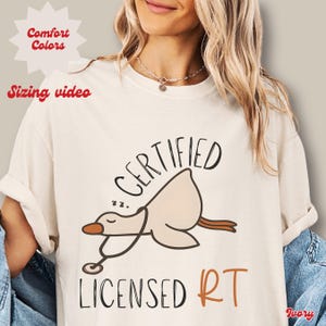 May include: Ivory-colored t-shirt with the words "CERTIFIED LICENSED RT" and a cartoon goose wearing a stethoscope. The goose is sleeping. The words "Comfort Colors" and "Sizing video" are in the upper left corner.