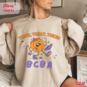 Op de afbeelding: Beige sweatshirt met een Halloween-thema. Het ontwerp toont een cartoon pompoen karakter met snoep, een vleermuis, en de tekst "TRICK, TREAT, TOKEN" en "BCBA" in oranje en paars. Een spin en sterren maken het ontwerp compleet.