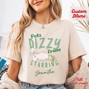 May include: A soft cream-coloured t-shirt featuring the text "Pots Dizzy Drama Starring Samantha" and a cartoon duck. The shirt has a relaxed fit and is made of soft cream-coloured fabric.