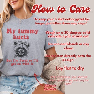 The Athletic Heather t-shirt with a raccoon illustration and the text 'my tummy hurts but I’m feral so I’ll get on with it'. The text 'How to Care' is at the top of the image. The text below the illustration reads 'Wash on a 30-degree cold delicate cycle inside out. Do use not bleach or oxy clean. Do not iron directly onto the design. Lay flat to dry. With a little love, your shirt will stay in great shape for longer.