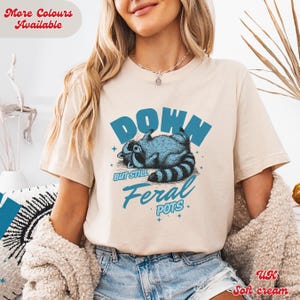 May include: A beige t-shirt featuring a blue raccoon graphic and the text "Down But Still Feral Pots".