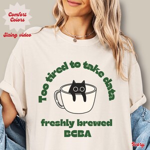 Puede incluir: Camiseta de color marfil con el texto verde "Too tired to take data", que rodea un gato negro en una taza de café blanca. Las palabras "freshly brewed BCBA" están debajo. La camiseta también tiene las palabras "Comfort Colors" y "Sizing video".