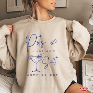 May include: A beige sweatshirt with the text "Pots Just add Salt Jennies Bar" in blue lettering. The design also includes a drawing of a martini glass with olives and a salt shaker.