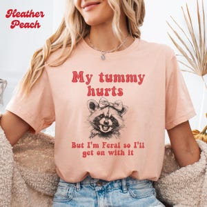 The heather peach unisex t-shirt with a smiling raccoon with a bow on her head graphic and the text 'My tummy hurts but I’m feral so I’ll get on with it ' in red groovy font