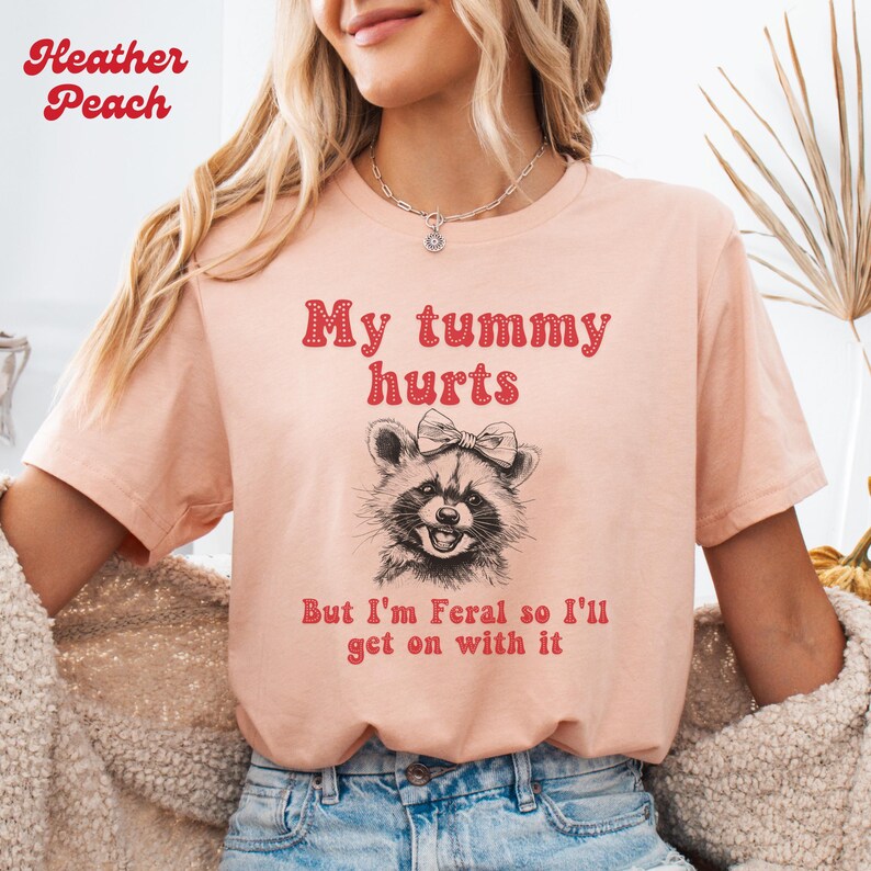 The heather peach unisex t-shirt with a smiling raccoon with a bow on her head graphic and the text 'My tummy hurts but I’m feral so I’ll get on with it ' in red groovy font