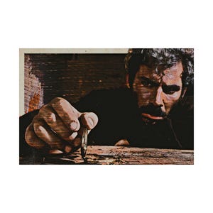 May include: A man with a beard and gray hair is holding a small, thin, metal object in his hand. He is looking down at the object, which is resting on a wooden surface.