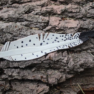 Puede incluir: Una pluma de cuero blanco con manchas negras y una punta negra. La pluma est&aacute; sobre un trozo de corteza de &aacute;rbol.