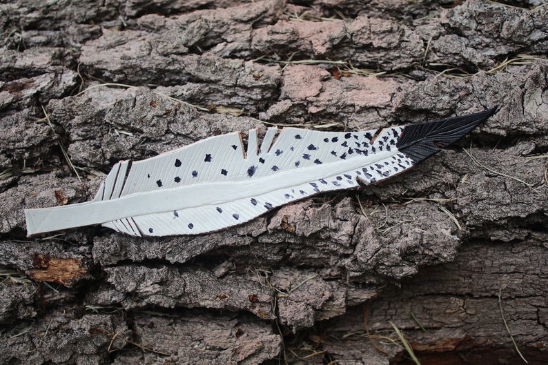 Puede incluir: Una pluma de cuero blanco con manchas negras y una punta negra. La pluma est&aacute; sobre un trozo de corteza de &aacute;rbol.