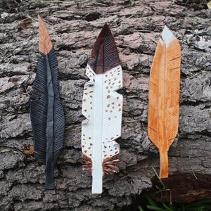 Puede incluir: Tres plumas de cuero, cada una con un color y un patr&oacute;n diferente. Las plumas son negras con puntas marrones, blancas con manchas marrones y negras, y marrones con rayas blancas.