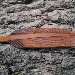 Puede incluir: Un marcap&aacute;ginas de cuero marr&oacute;n en forma de pluma con un dise&ntilde;o detallado. El marcap&aacute;ginas est&aacute; sobre una superficie rugosa y texturizada.
