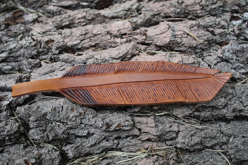 Puede incluir: Un marcap&aacute;ginas de cuero marr&oacute;n en forma de pluma con un dise&ntilde;o detallado. El marcap&aacute;ginas est&aacute; sobre una superficie rugosa y texturizada.