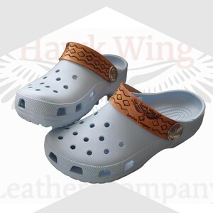 Puede incluir: Zuecos azul claro con correas de cuero marrón. Las correas presentan patrones geométricos y un diseño de águila estampado. Los zuecos tienen múltiples orificios de ventilación y un botón plateado. La marca "Hawk Wing Leather Company" es visible.