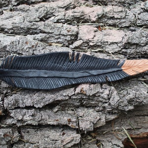 Puede incluir: Una pluma de cuero negro con una punta marr&oacute;n, descansando sobre un trozo de corteza de &aacute;rbol.