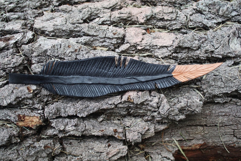 Puede incluir: Una pluma de cuero negro con una punta marr&oacute;n, descansando sobre un trozo de corteza de &aacute;rbol.