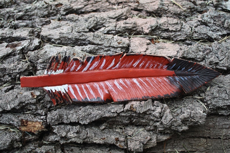 Puede incluir: Una pluma de cuero roja, blanca y negra con un dise&ntilde;o pintado. La pluma est&aacute; sobre un trozo de corteza de &aacute;rbol.