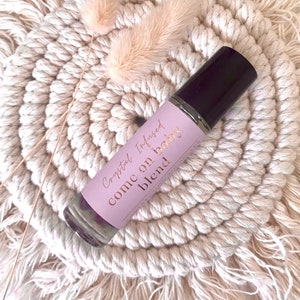Op de afbeelding: Een transparante glazen rollerfles met een zwarte dop en een roze-gouden etiket. Op het etiket staat "Crystal Infused come on baby blend".