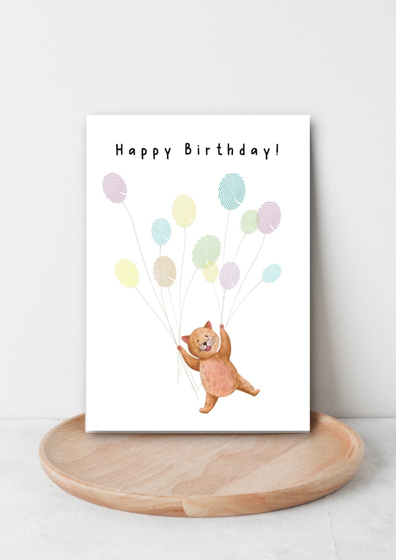 K&ouml;nnte beinhalten: Eine Geburtstagskarte mit einer braunen Katze, die an bunten Luftballons festh&auml;lt. Die Ballons sind aus Fingerabdruckmustern gefertigt und die Karte lautet "Happy Birthday!"